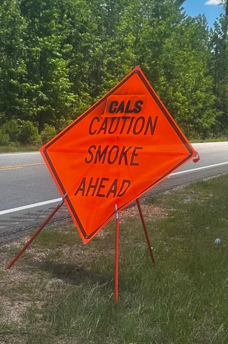 Bright orange diamond-shaped warning sign reading Cals Caution Smoke Ahead posted on a roadside with trees and blue sky in background